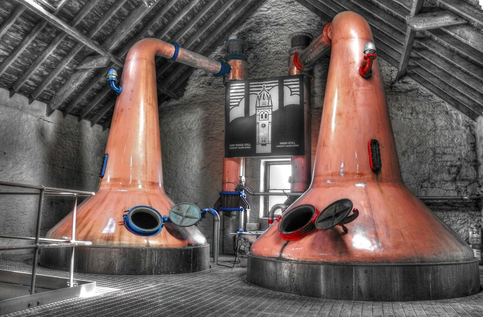 Dos grandes alambiques de cobre de una destilería tradicional, bajo un techo de vigas de madera oscura